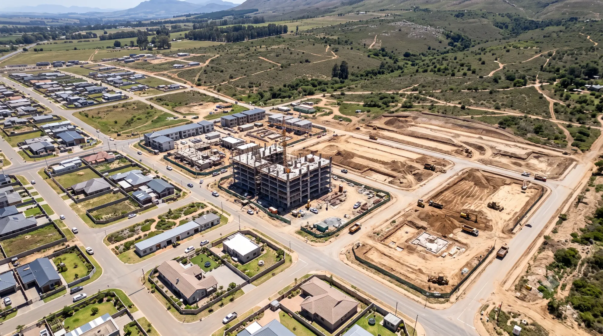 Aerial view of a peri-urban enforcement perimeter: planned housing on the left, an active mid-rise construction site in the centre, open parcels under earthwork on the right, with mountains in the distance